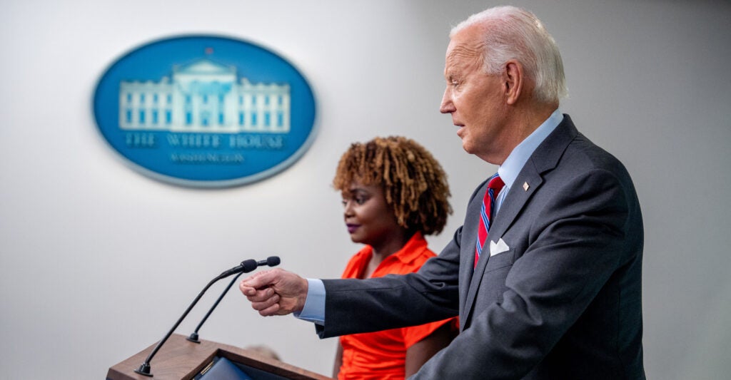 President Joe Biden, wearing a gray suit and a red-striped tie, speaks during a White House press briefing as he stands beside White House press secretary Karine Jean-Pierre, who is wearing a bright orange blouse.