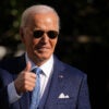 President Joe Biden, wearing dark aviator sunglasses and a navy blue suit, gives a thumbs-up to the audience after pardoning the National Thanksgiving Turkey.