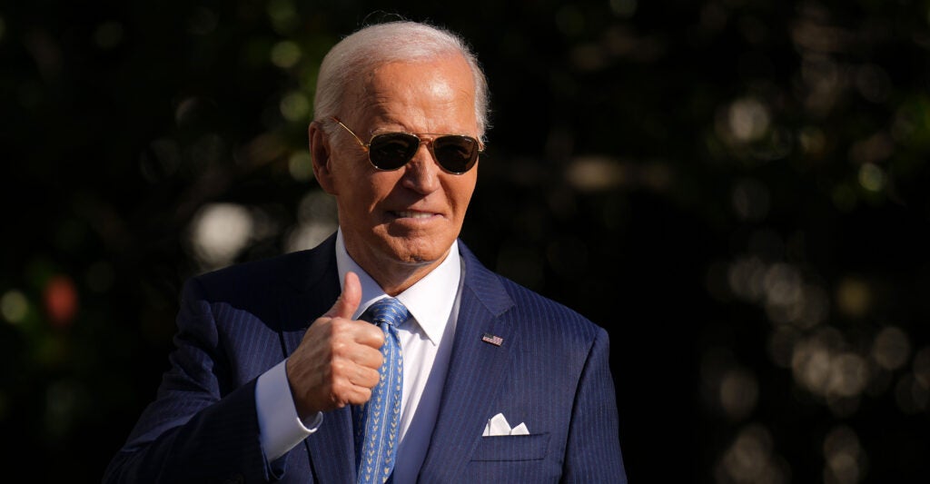 President Joe Biden, wearing dark aviator sunglasses and a navy blue suit, gives a thumbs-up to the audience after pardoning the National Thanksgiving Turkey.