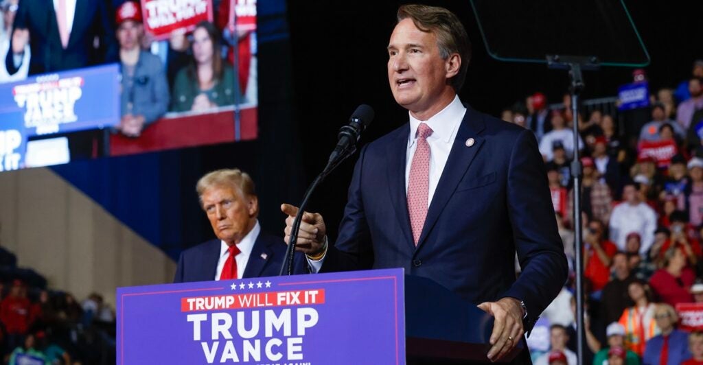 Virginia Gov. Glenn Youngkin talks at a podium with Donald Trump in the background.