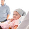 A young girl lies in a hospital bed holding a teddy bear, her parents watching, concerned, in the background.