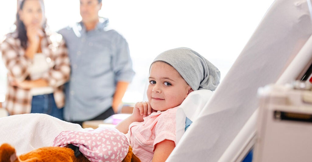 A young girl lies in a hospital bed holding a teddy bear, her parents watching, concerned, in the background.