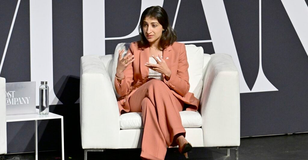 Lina Khan sits in a sofa couch seat with her legs crossed while speaking.