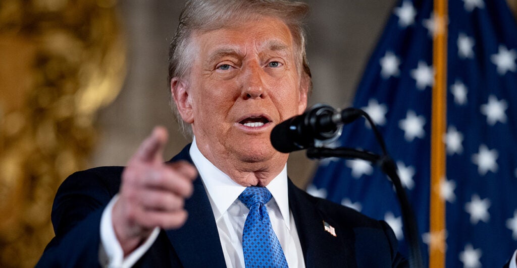 Donald Trump in a dark suit and light blue tie speaking at a microphone and pointing to the audience. An American flag is in the background.