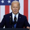 President Joe Biden squints as he stands behind a podium in a navy blue suit, the American flag in the background.