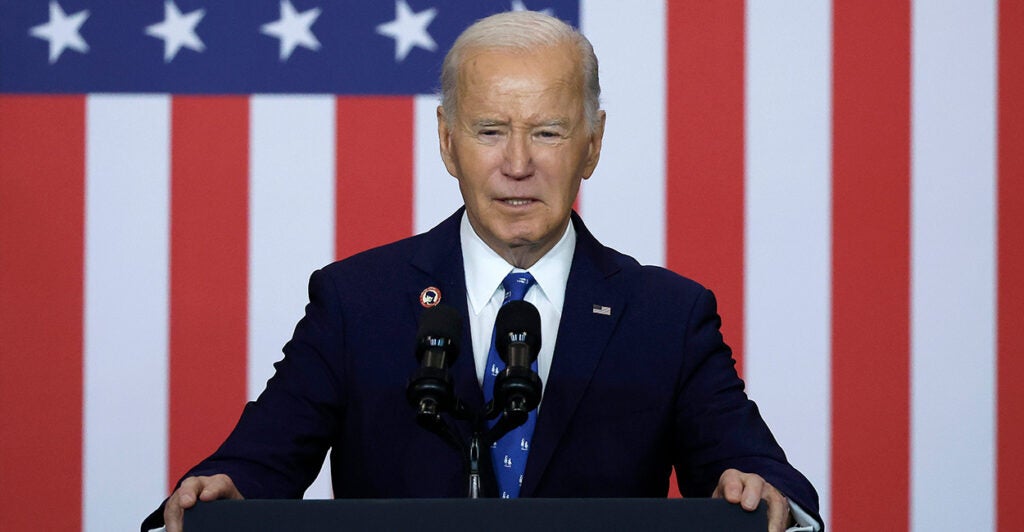 President Joe Biden squints as he stands behind a podium in a navy blue suit, the American flag in the background.