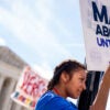 A young woman stands on a lamppost and holds a sign that reads "MAKE ABORTION UNTHINKABLE" in front of the Supreme Court.