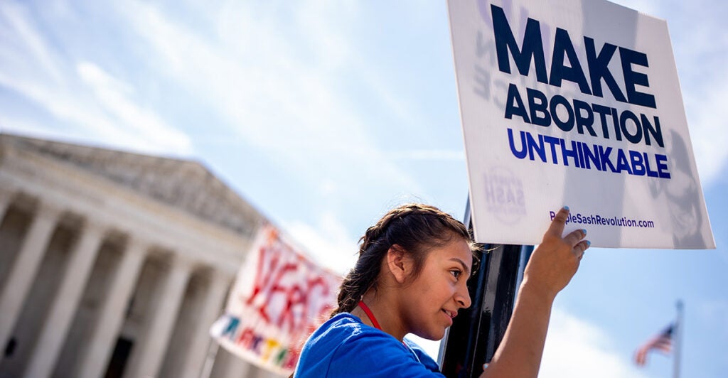 A young woman stands on a lamppost and holds a sign that reads "MAKE ABORTION UNTHINKABLE" in front of the Supreme Court.