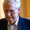 Mitch McConnell, in a blue suit, walks down an orange Senate hallway, with a frown on his face.