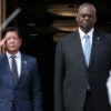 Filipino President Ferdinand Marcos stands with U.S. Secretary of Defense Lloyd Austin, both in dark suits standing in a large doorway flanked by two U.S. Navy guards with rifles and bayonets.