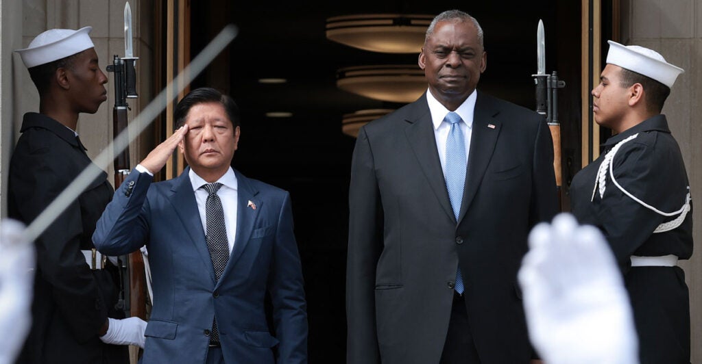 Filipino President Ferdinand Marcos stands with U.S. Secretary of Defense Lloyd Austin, both in dark suits standing in a large doorway flanked by two U.S. Navy guards with rifles and bayonets.