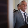 RFK Jr., in a gray suit, walks in a Senate Office hallway.