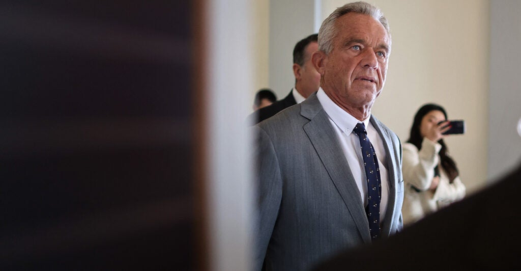 RFK Jr., in a gray suit, walks in a Senate Office hallway.