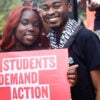 Two students smile and hold a sign that reads, "Students Demand Action."