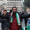 A vendor selling scarves colored in the independence-era Syrian flag in Syria, smiling and draped in the scarves with outstretched arms to show them off.