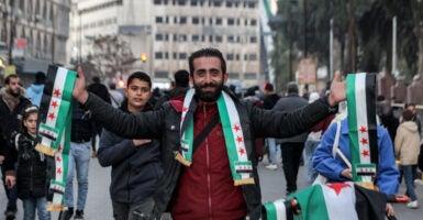 A vendor selling scarves colored in the independence-era Syrian flag in Syria, smiling and draped in the scarves with outstretched arms to show them off.