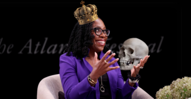 Supreme Court Justice Ketanji Brown Jackson wears a purple blazer and smiles with her hands up. She is superimposed wearing a crown and holding a skull.