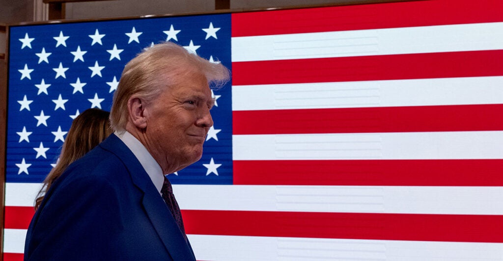 Donald Trump walks with Melania in front of a screen displaying with the United States flag.