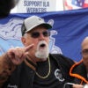 Longshoremen's union leader Harold Daggett in a black T-shirt and jacket with sunglasses and a baseball cap gestures by pointing his finger while talking to striking dockworkers