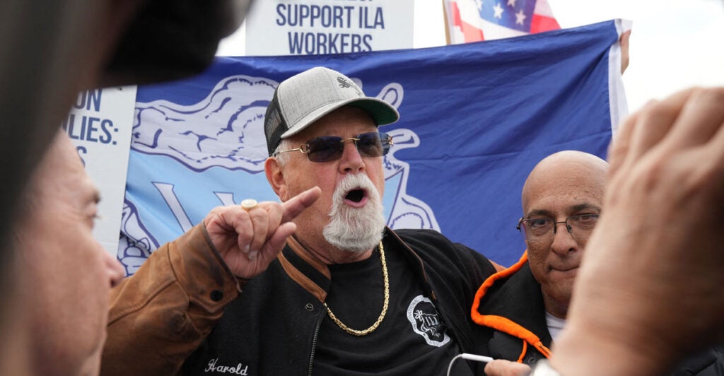 Longshoremen's union leader Harold Daggett in a black T-shirt and jacket with sunglasses and a baseball cap gestures by pointing his finger while talking to striking dockworkers