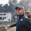 Gavin Newsom in a ball cap stands in front of a fire