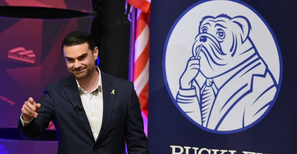 Ben Shapiro stans at a podium in front of an American flag.