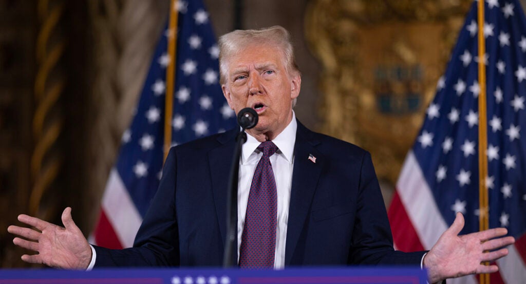 Donald Trump gestures in a suit in front of two American flags