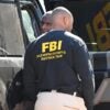 A man wearing an FBI jacket stands near a vehicle.