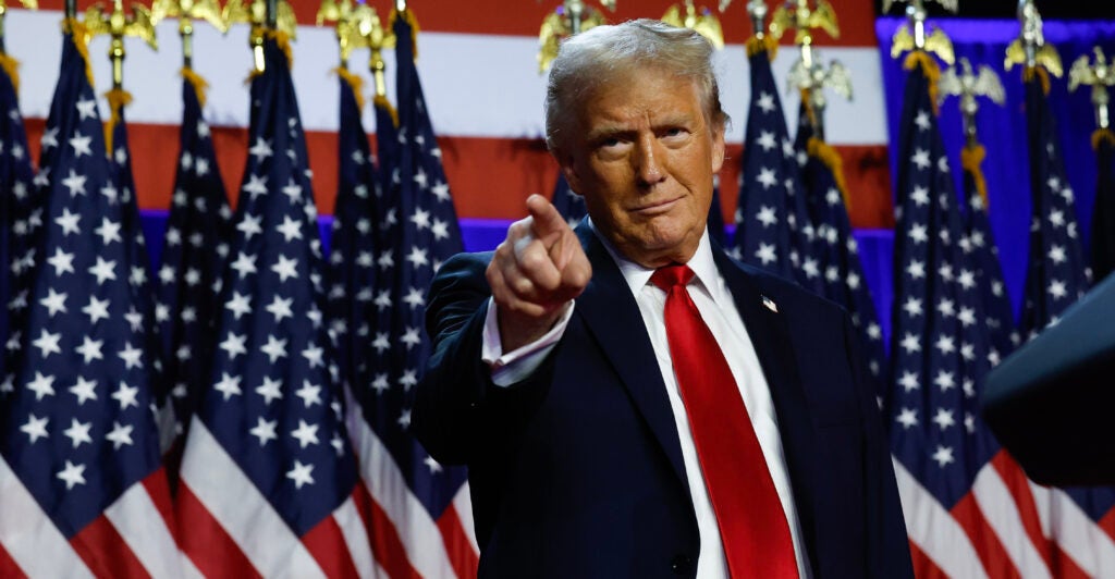 Donald Trump, wearing a black suit and red necktie, points to the crowd at a campaign event.