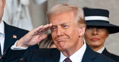 Donald Trump, in a red and blue necktie and black suit, smiles as he salutes alongside Melanie Trump, who wears a navy wide-brimmed hat.