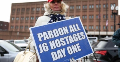 Summer Boger wears a white winter coat and navy scarf with white stars as she holds a sign that reads, ""Pardon all J6 hostages day one."