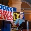 A man wearing a hat holds a sign that states, "Guns Save Lives."