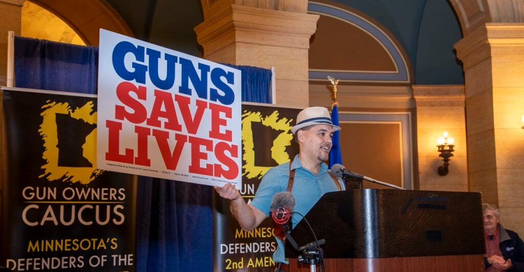 A man wearing a hat holds a sign that states, "Guns Save Lives."