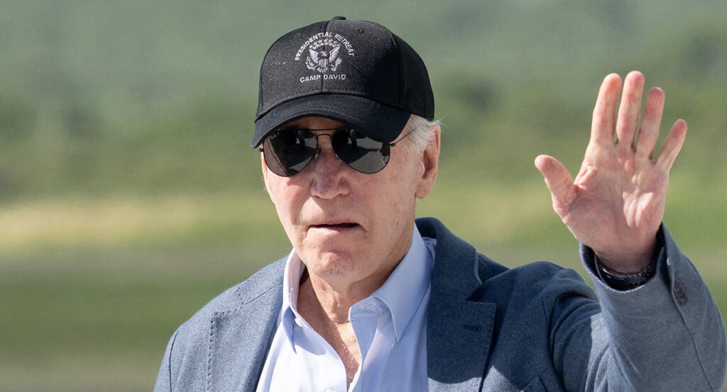 Joe Biden in a blue shirt and a grey sport coat waves in a hat reading "Presidential Retreat Camp David"