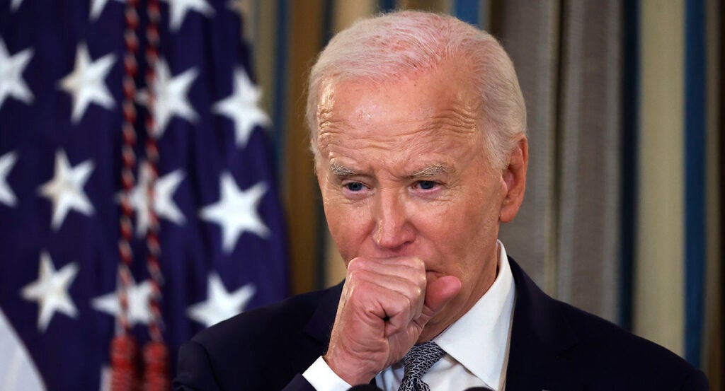 Joe Biden in a black suit puts his hand in front of his mouth