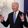 Joe Biden in a black suit with a blue tie and American flag pin raises his hands