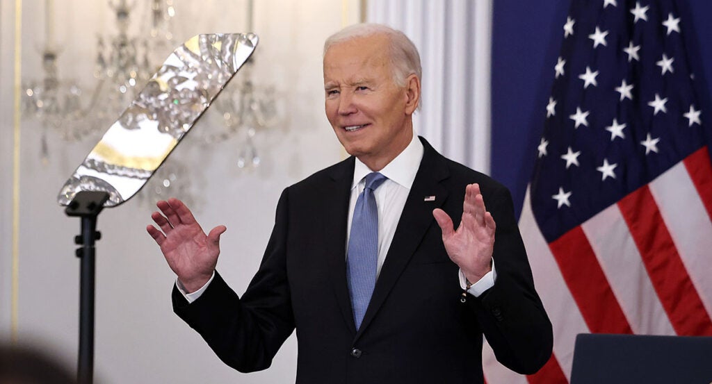 Joe Biden in a black suit with a blue tie and American flag pin raises his hands