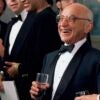 Milton Friedman holds a drink in his right hand while wearing a tuxedo at a dinner.