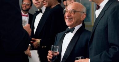 Milton Friedman holds a drink in his right hand while wearing a tuxedo at a dinner.