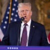 Donald Trump in a dark suit and tie speaking at a podium with a microphone with two American flags behind him