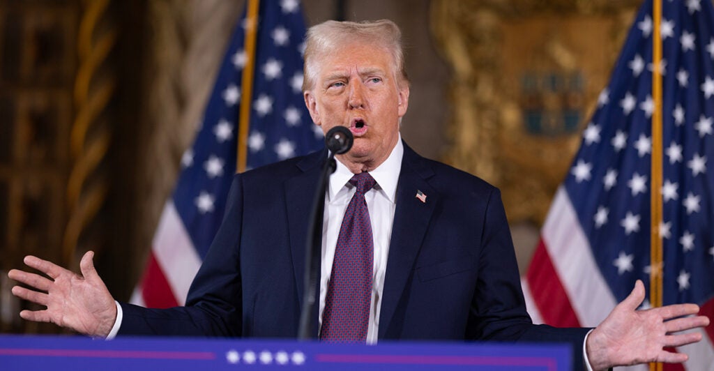 Donald Trump in a dark suit and tie speaking at a podium with a microphone with two American flags behind him