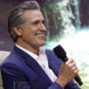 California Governor Gavin Newsom in a dark blue suit with no tie, sitting on stage holding a microphone