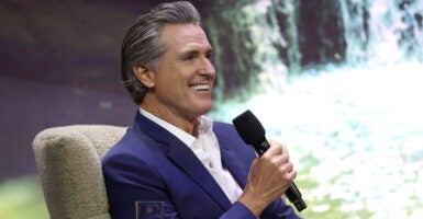 California Governor Gavin Newsom in a dark blue suit with no tie, sitting on stage holding a microphone