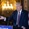 Donald Trump gestures in a suit in front of an American flag