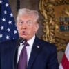 Donald Trump in a blue suit speaks in front of an American flag