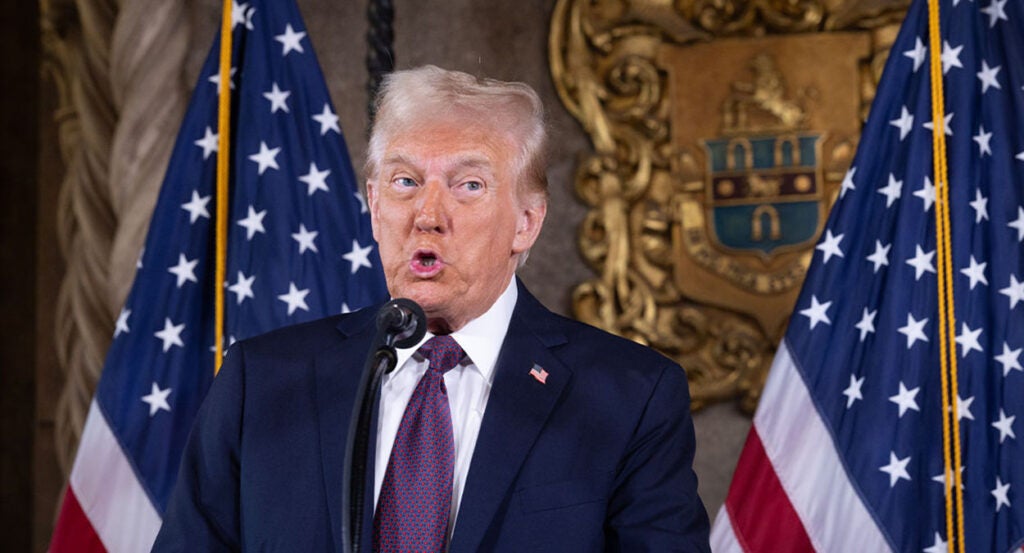 Donald Trump in a blue suit speaks in front of an American flag