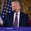 Donald Trump stands at a podium with his arms held out with American flags in the background.