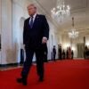 Donald Trump walks with his hands to his side in the White House on a red carpet.