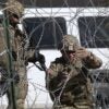 One soldier kneels while cutting barb wire while another soldier holding a rifle stands nearby.