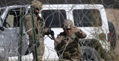 One soldier kneels while cutting barb wire while another soldier holding a rifle stands nearby.